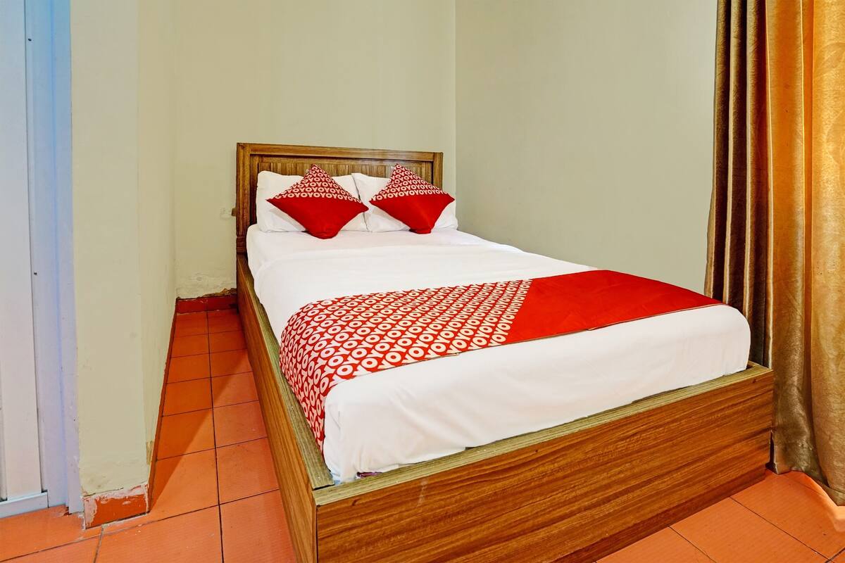 A single bed with a wooden frame, white bedding, and red patterned pillows.