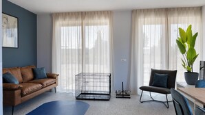 A modern living room with a brown leather sofa, a blue rug, a metal pet cage, a dining table with chairs, and a large green plant. - Spacious apartment for 4 people, with various facilities for your dog (Nieuwvliet-Bad)