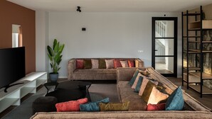 Ein Wohnzimmer mit Ecksofa, einem schwarzen Couchtisch, einer großen Kübelpflanze, einem Fernseher und einem Bücherregal.