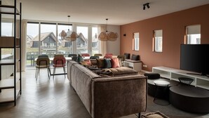 Ein modernes Wohnzimmer mit einer Couch, Sesseln, einem Fernseher und einem großen Fenster mit Blick auf eine Wohngegend.