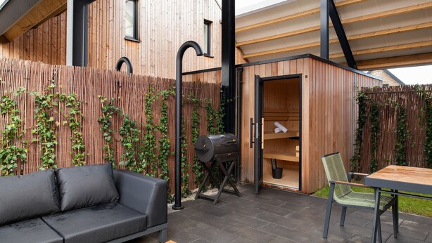 Une cabane en bois dotée d’un sauna intégré, d’un barbecue noir, d’un canapé et d’une table avec des chaises.