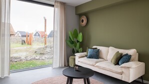 Un salon avec une grande fenêtre, un canapé beige agrémenté de coussins, une table basse ronde noire et une plante verte.