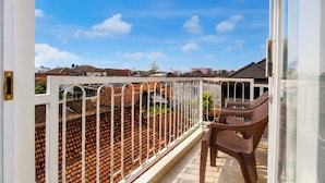 Un balcón con barandilla de metal blanco, una silla de mimbre marrón y vista a una zona residencial con casas y cielo despejado.