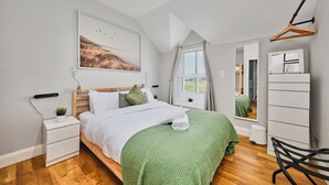 A bedroom with a bed, a bedside table, a nightstand, a mirror, a hanging light, a window with curtains, and a view of the outdoors. - Dunluce Park - Portrush by the Sea (Portrush)