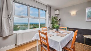 A dining area with a table set for four, a view of the sea through large windows, a radiator, and a plant on the windowsill. - Dunluce Park - Portrush by the Sea (Portrush)