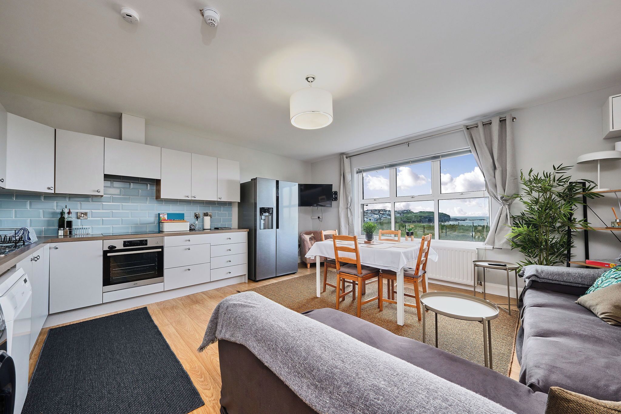 A modern kitchen with white cabinets, a built-in oven, and a refrigerator. There is a dining area with a table and chairs, and a large window offering a view of the outdoors.