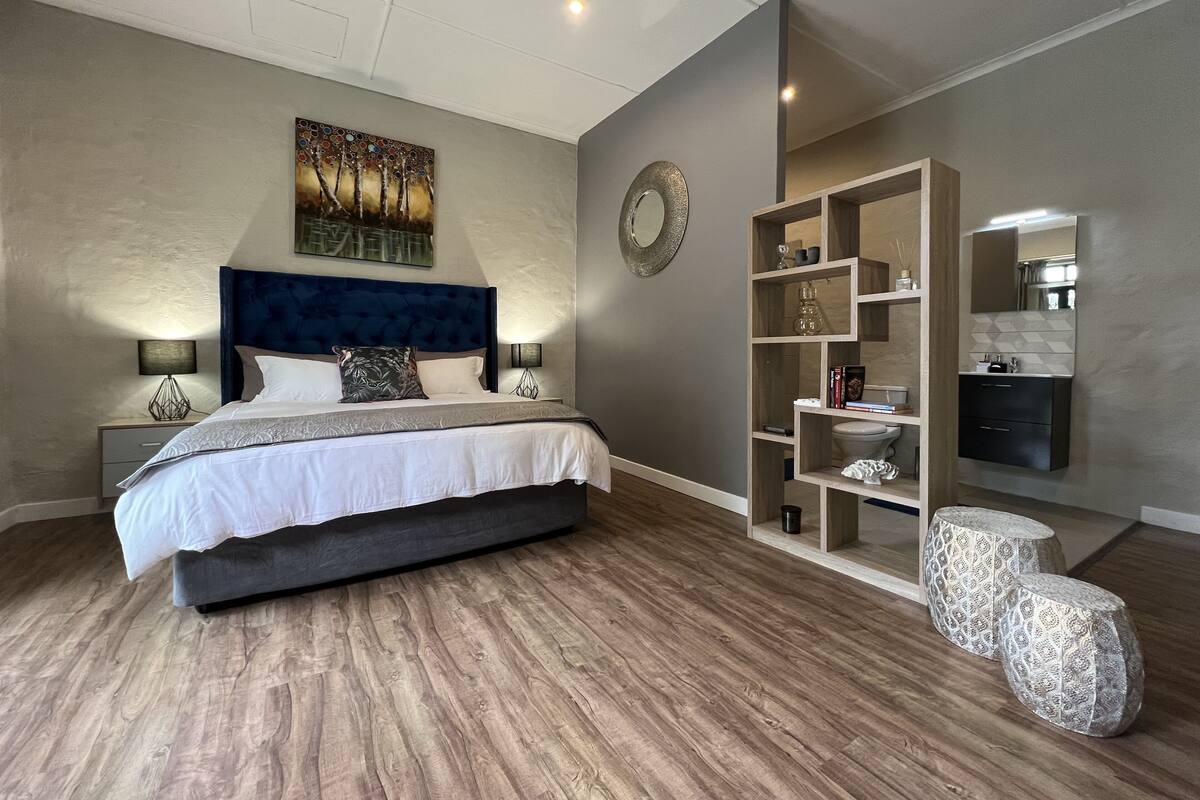 Un dormitorio moderno con una cama grande, cabecera, un cuadro, un espejo, un mueble de estanterías empotrado y un taburete pequeño.