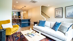 Una sala moderna con un sofá, una mesa de centro y un sillón amarillo. Se ve una escalera que conduce a un nivel superior, y un área de cocina al fondo.