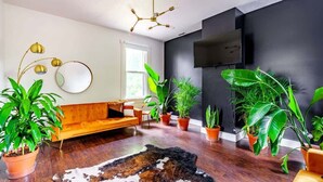 A modern living room with a wooden floor, a sofa, a TV mounted on the wall, and several potted plants. - Downtown Chillicothe | Walk to Historic District | Free Parking | 5,800+ Reviews (Chillicothe)