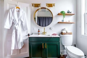A bathroom with a white robe hanging, a round mirror, a green vanity, a white toilet, and a window. - Downtown Chillicothe | Walk to Historic District | Free Parking | 5,800+ Reviews (Chillicothe)