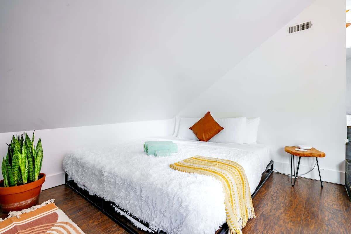 Ein Schlafzimmer mit einem weißen Bett, einer Topfpflanze, einem kleinen Holzschemel und einer weißen, flauschigen Decke.
