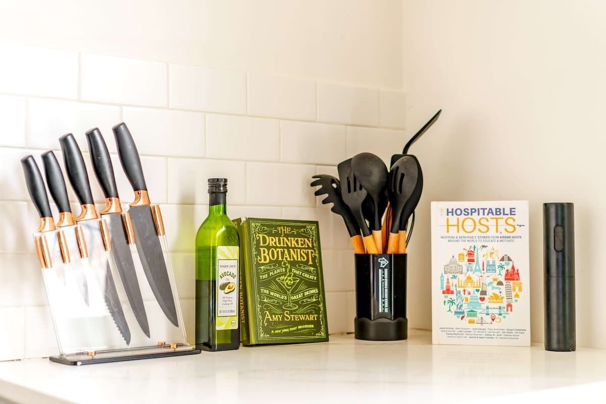 A kitchen counter with knives, a bottle of olive oil, a book titled 'The Drunken Botanist', a set of kitchen utensils, and a book titled 'Hospitality Hosts'.