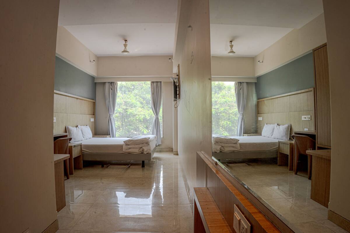 A hotel room with two beds, a large mirror, a wooden desk, and a view of greenery outside.