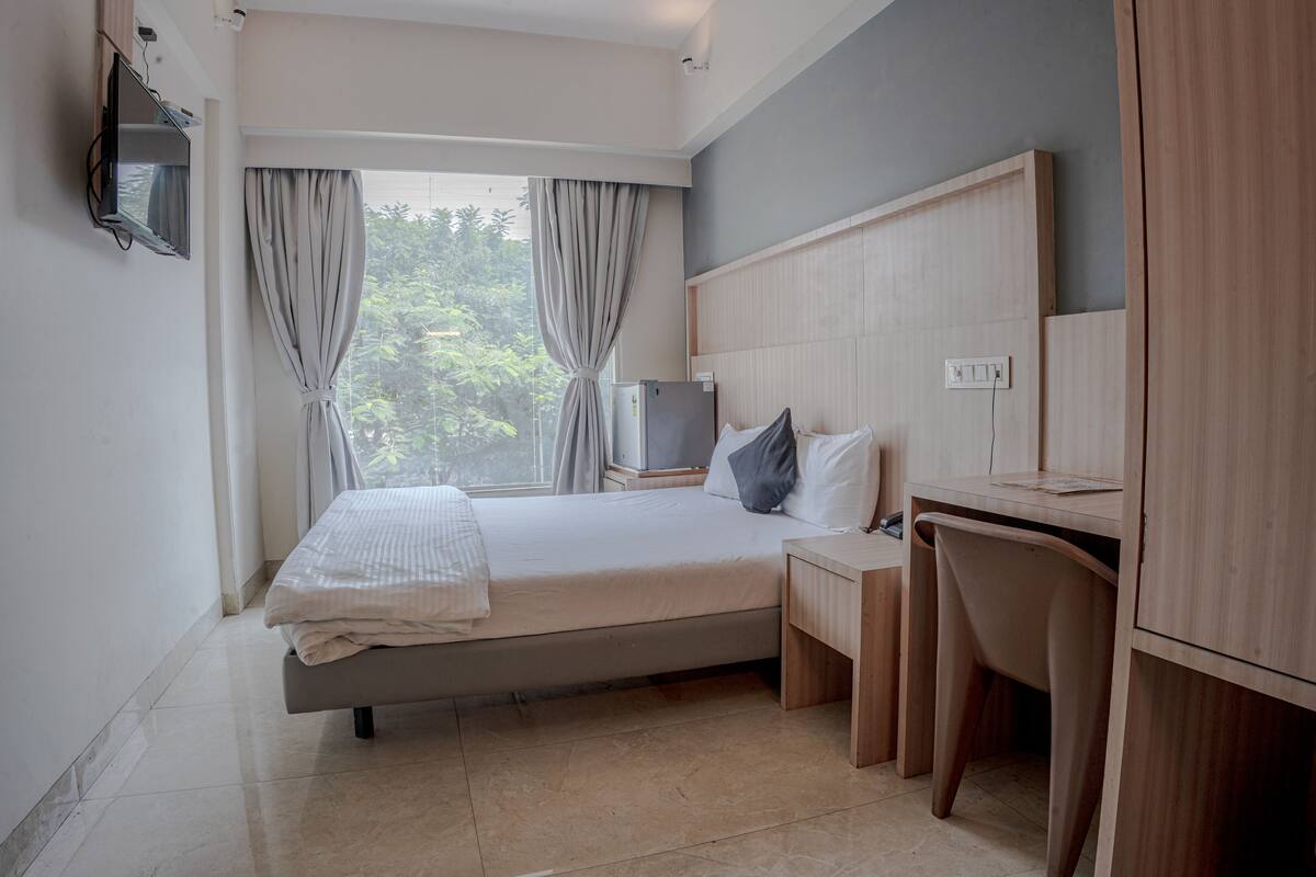 A bedroom with a bed, a desk, a chair, a TV, and a window with curtains.