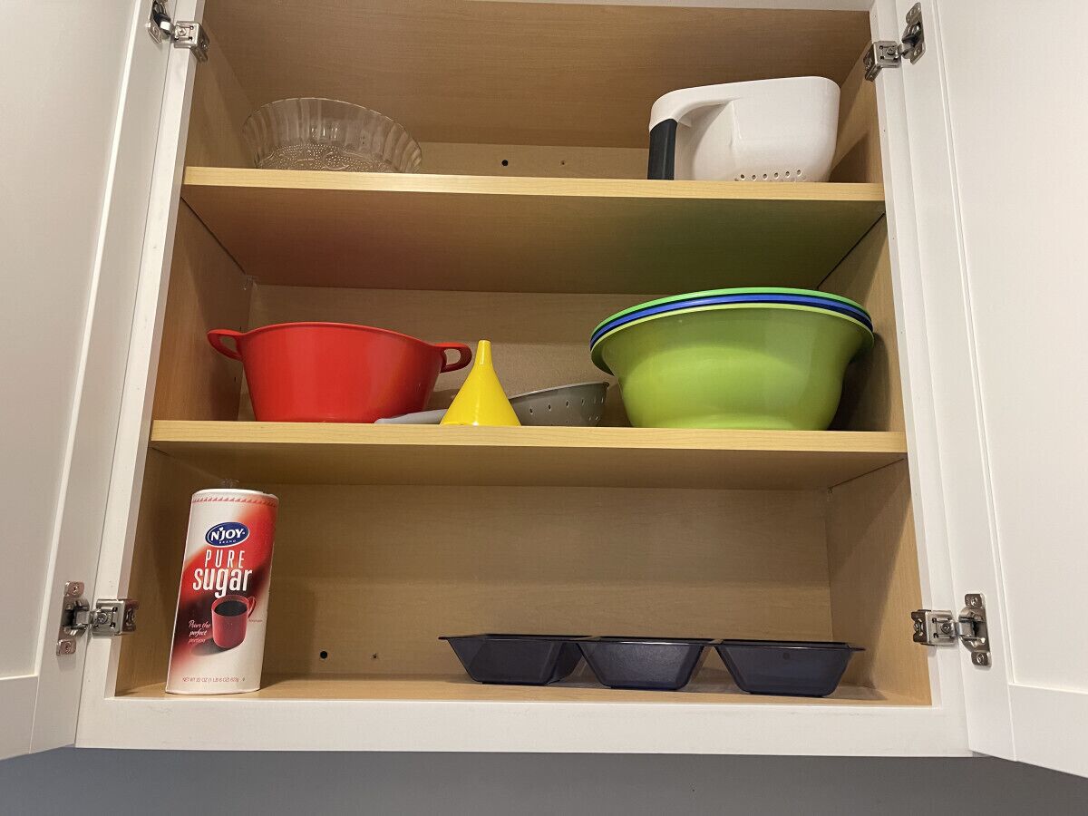 Um armário com prateleiras contendo diversos utensílios de cozinha, incluindo uma lata de Njoy Pure Sugar, uma panela vermelha, um funil amarelo, uma tigela verde e um recipiente branco.