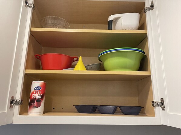 A cabinet with shelves containing various kitchen items, including a can of Njoy Pure Sugar, a red pot, a yellow funnel, a green bowl, and a white container.