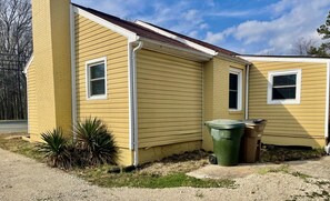 Ett gult, enplanshus med ett garage, en liten trädgård framför huset med några växter och två soptunnor.