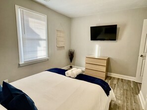 A bedroom with a bed, a dresser, a flat-screen TV, and a window with blinds. - Vibrant 3BR Ranch | Prime Greensboro Location (Greensboro)