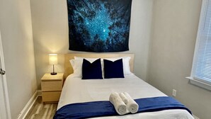Un lit bien fait, avec une literie blanche et bleu marine, un plaid bleu et deux serviettes enroulées. Une table de chevet avec une lampe se trouve à côté du lit.