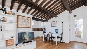 Een moderne woonkamer met een houten plafond, een flatscreen televisie, witte planken, een eethoek met stoelen en een keuken op de achtergrond.