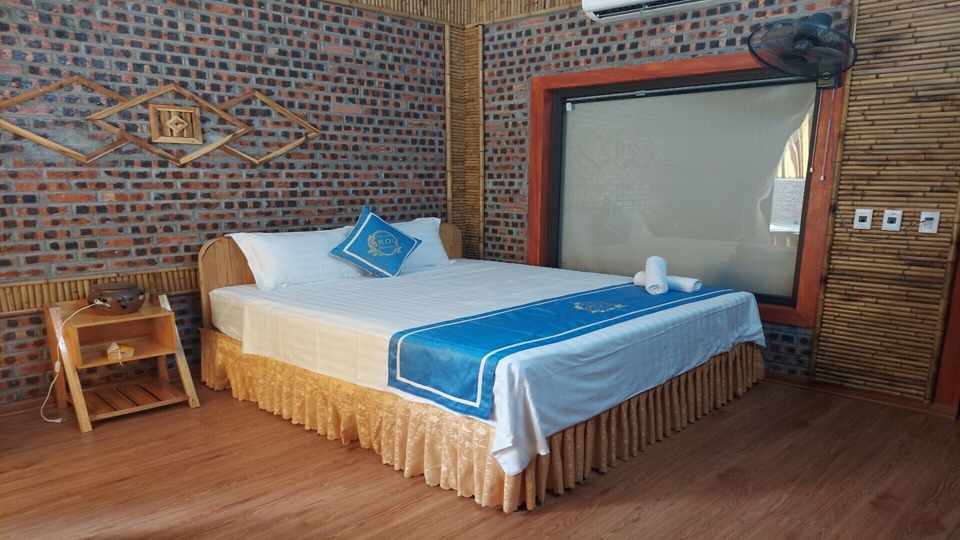 Een hotelkamer met een wand in baksteenmotief, een groot raam, een houten bed met een blauwe spreideken en een klein houten nachtkastje met een lamp.