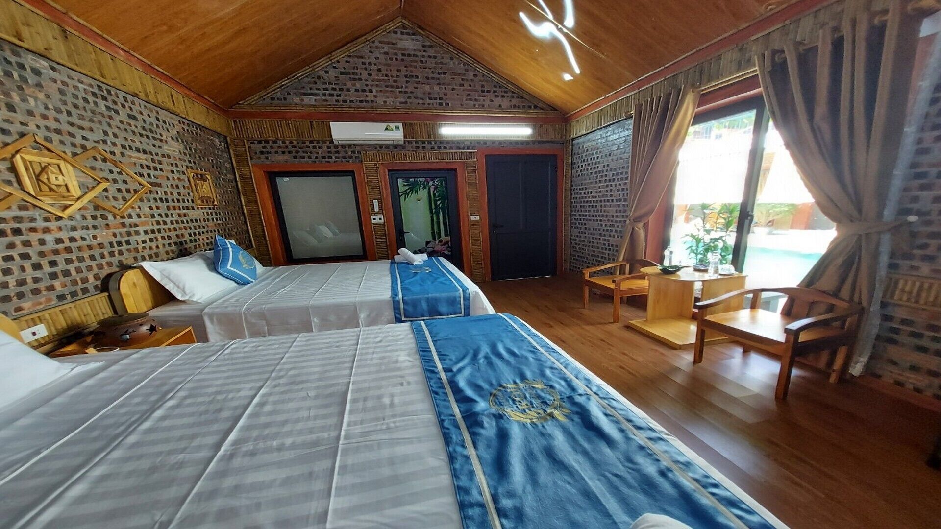 A room with two beds, a wooden ceiling, a brick wall, a small table, and a chair.