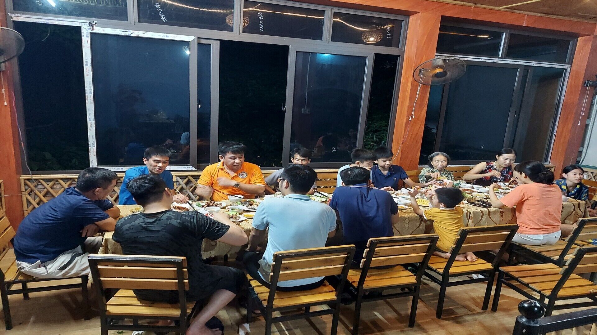 A group of people dining at a wooden table with chairs, in a room with large windows and a fan on the wall.
