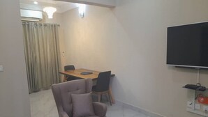 Een moderne woonkamer met een bank, een ronde houten tafel, een aan de muur bevestigde televisie en een eethoek met tafel en stoelen.