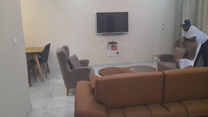 Een moderne woonkamer met een bruine bank, een flatscreen televisie aan de muur en een eethoek met een tafel en stoelen.
