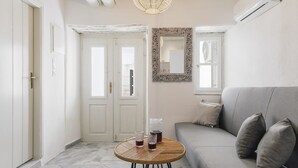 Une pièce avec un canapé gris, une table ronde en bois sur laquelle reposent deux verres et une carafe, un miroir accroché au mur et une porte dotée d’un cadre blanc.