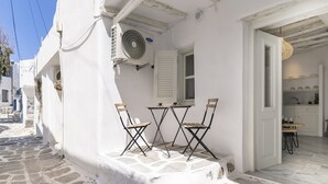 Un bâtiment blanc avec une rue pavée de pierres, un petit espace extérieur aménagé avec une table et des chaises, et une cuisine visible avec des placards et un évier.
