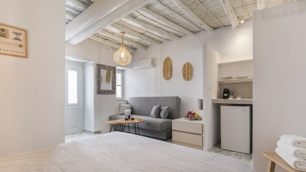 Une pièce moderne et bien éclairée, avec un canapé gris, une petite table ronde sur laquelle sont posées des boissons, un lit blanc et une petite cuisine équipée.