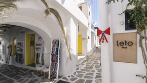Une rue étroite bordée de bâtiments blancs, avec une porte jaune et une enseigne indiquant « Leto Paros ».