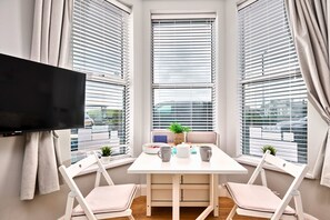 Una habitación moderna con una mesa blanca, dos sillas, un televisor de pantalla plana y ventanas grandes con persianas.