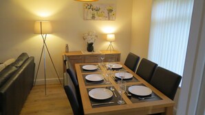 Un comedor con una mesa de madera preparada para seis personas, flanqueada por sillas de cuero negro. Hay dos lámparas de pie a cada lado de la mesa, y un cuadro de flores cuelga en la pared sobre ella.