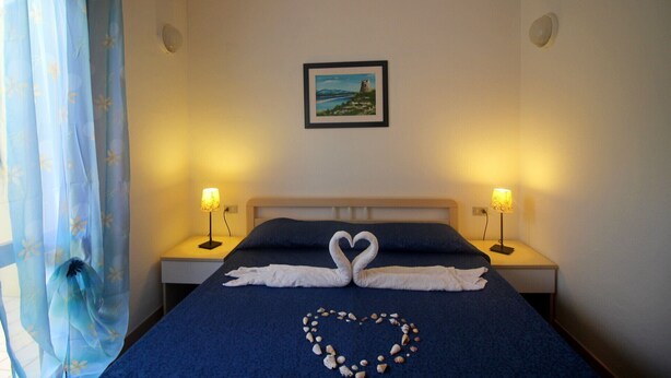 Een hotelkamer met een bed, twee bedlampen, een foto in een lijst aan de muur en een hartvormige decoratie van schelpen op het bed.