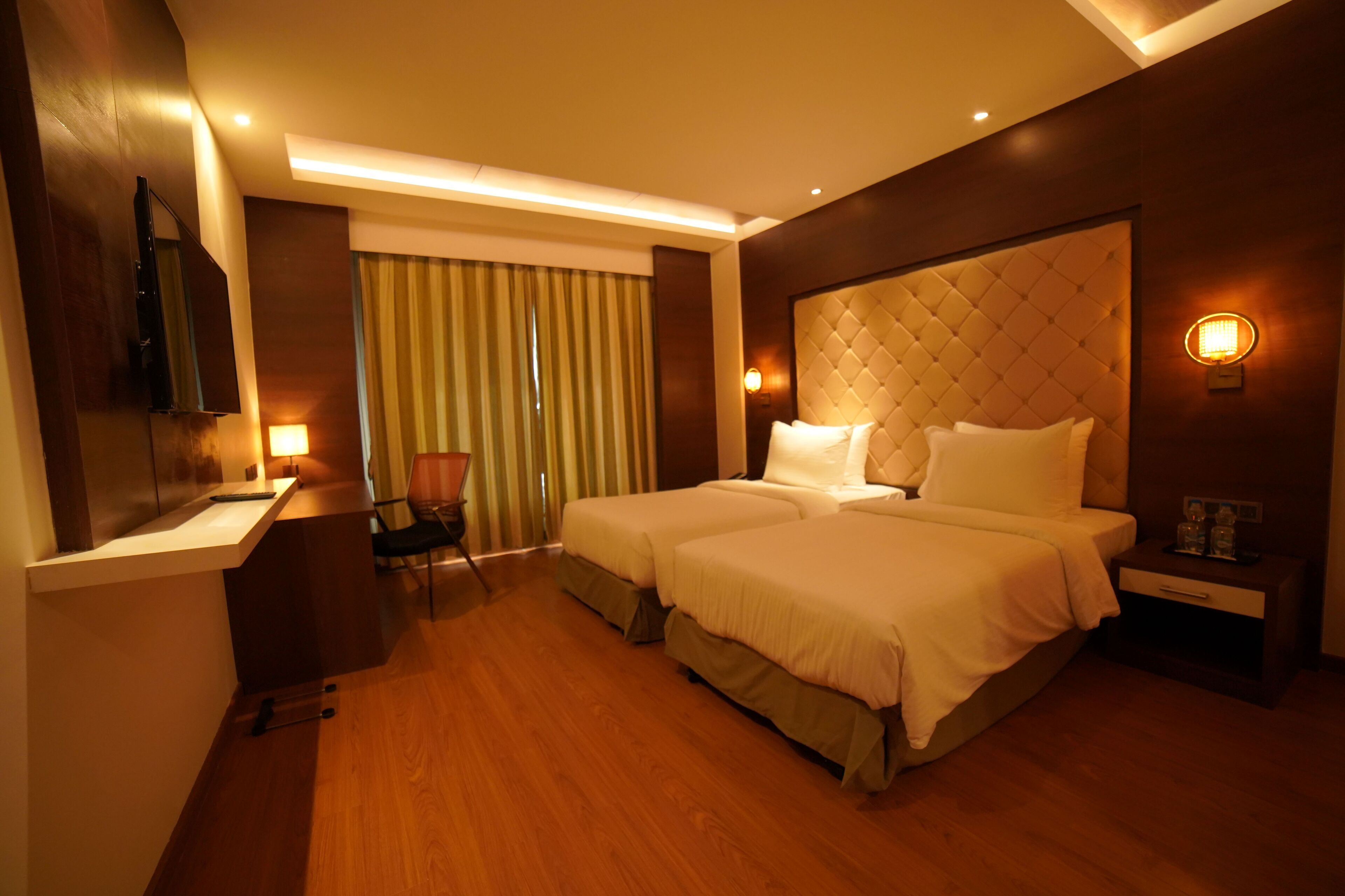 A hotel room with two beds, a desk, a chair, a TV mounted on the wall, and a window with curtains.