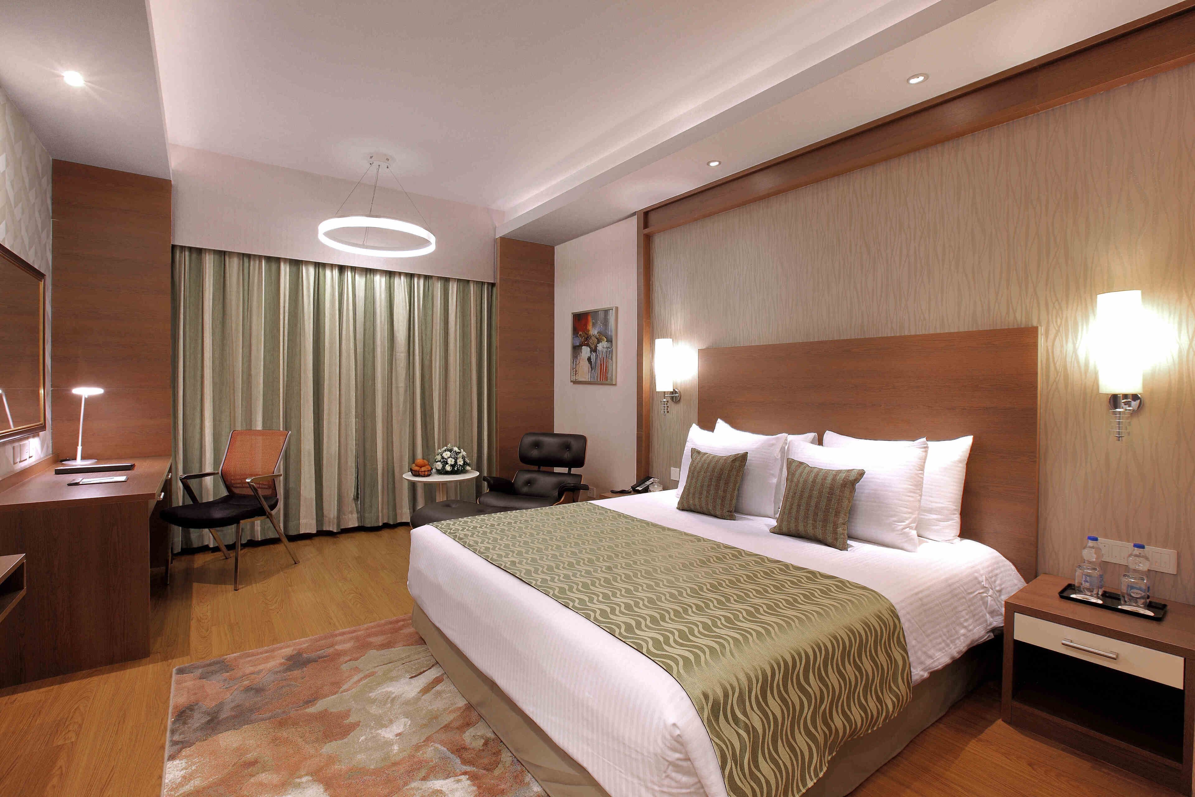 A hotel room with a large bed, a desk, a chair, a small table, and a nightstand with a lamp.