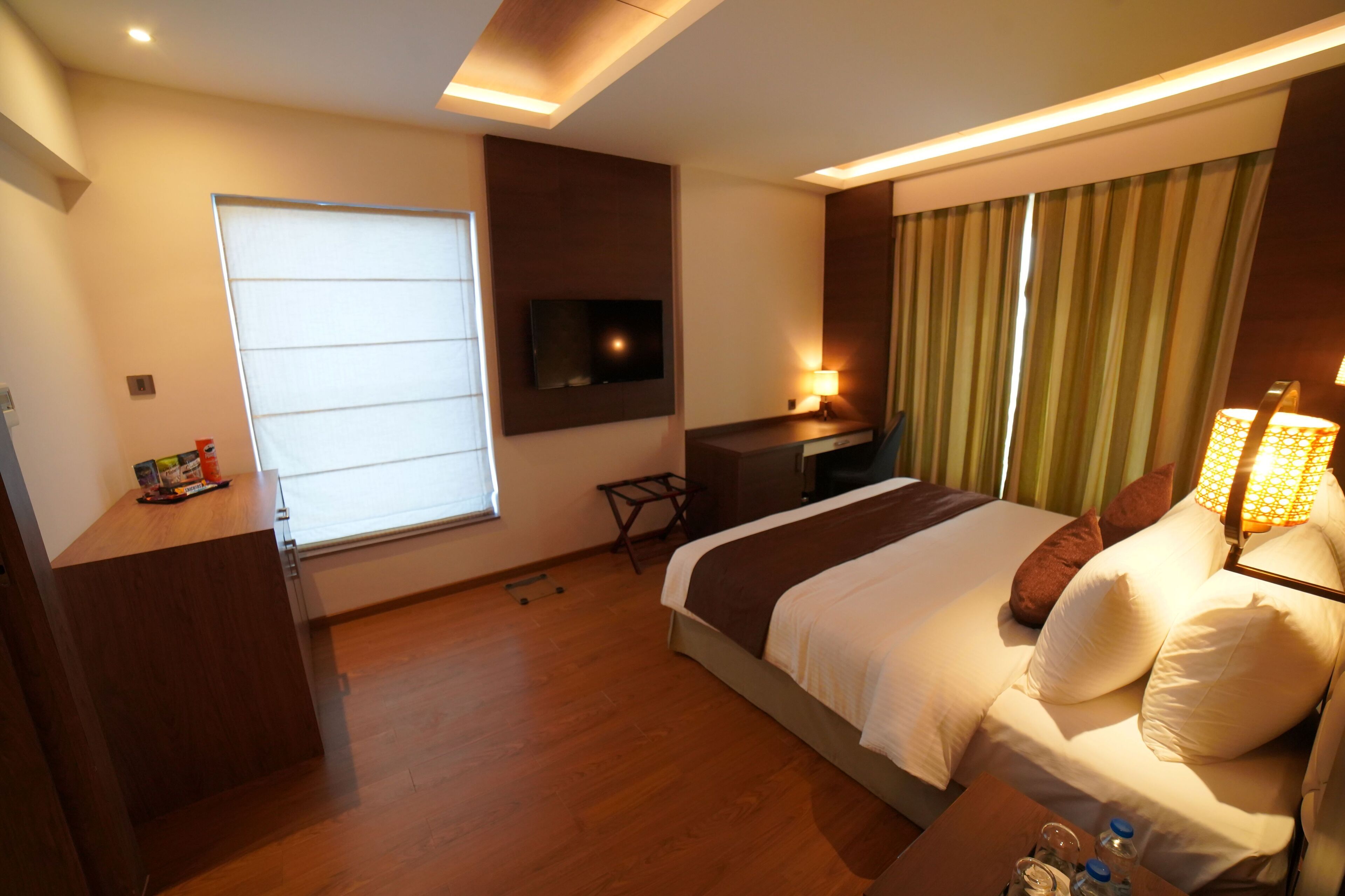 A hotel room with a bed, a desk, a TV, a window with blinds, and a lamp.
