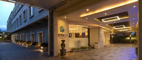 Una entrada de hotel moderna con una recepción bien iluminada, un logotipo visible y un camino pavimentado que conduce a la entrada.