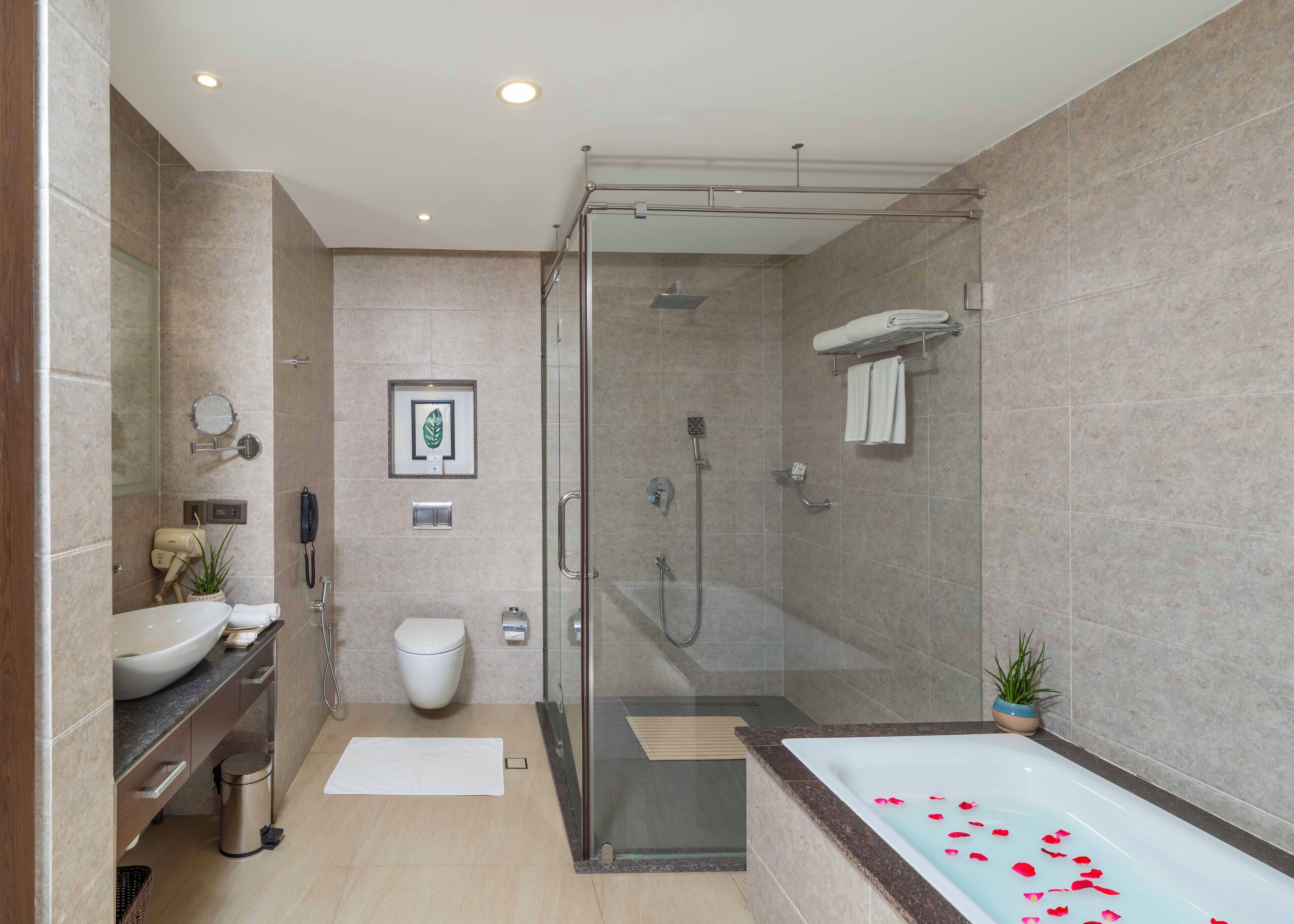 Un baño moderno con ducha de vidrio, una bañera grande con pétalos de rosa, un inodoro y un tocador con lavamanos y toallero.