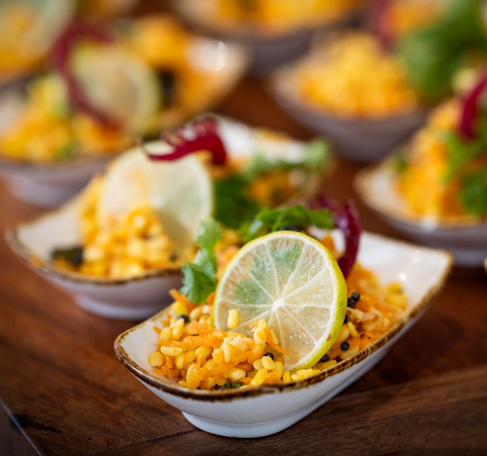 Une série de bols contenant un plat à base de maïs, de citron vert et de légumes verts, servis sur une table en bois.
