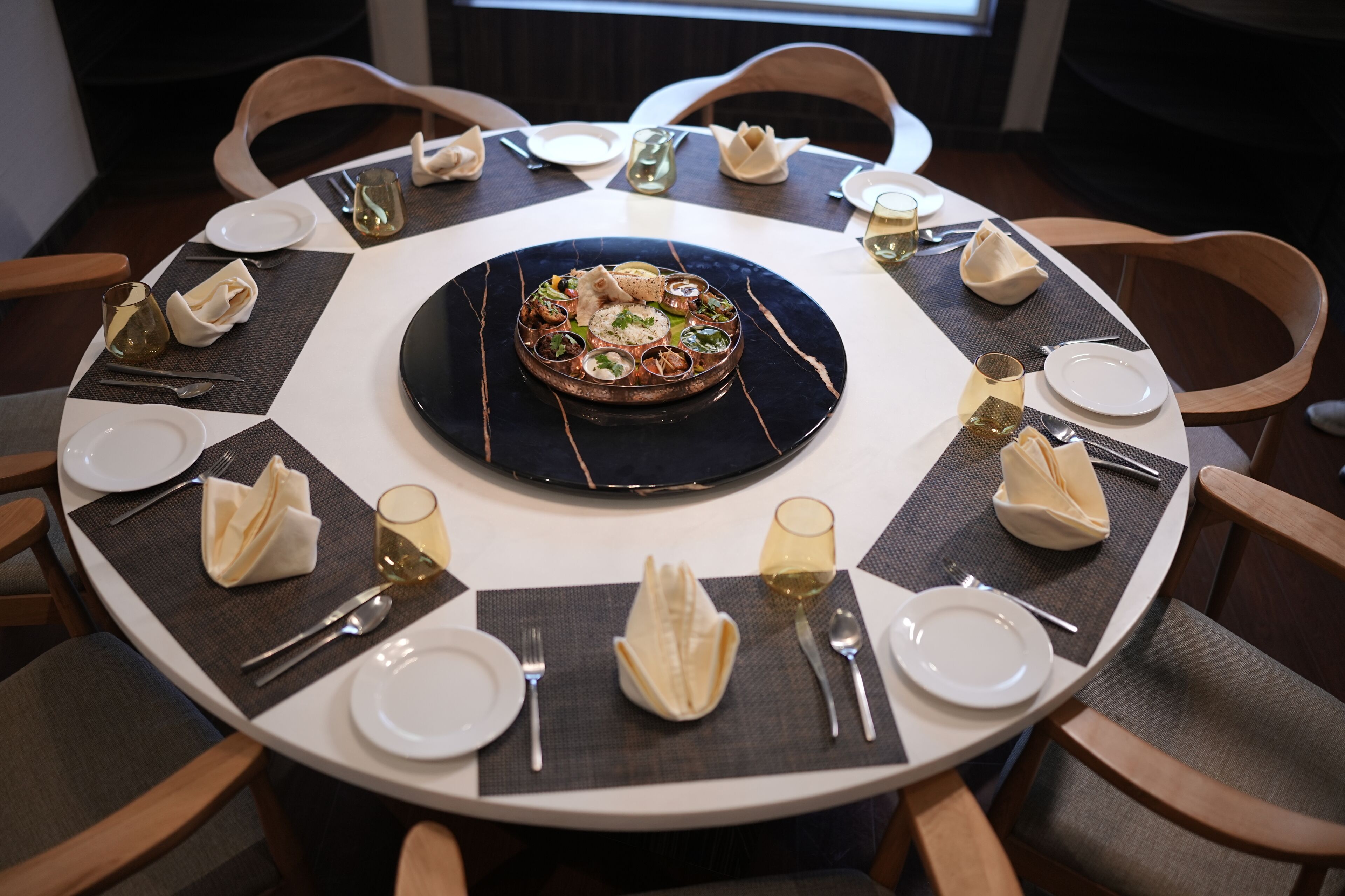 A round dining table with a central dish, surrounded by chairs and set with plates, cutlery, and glasses.