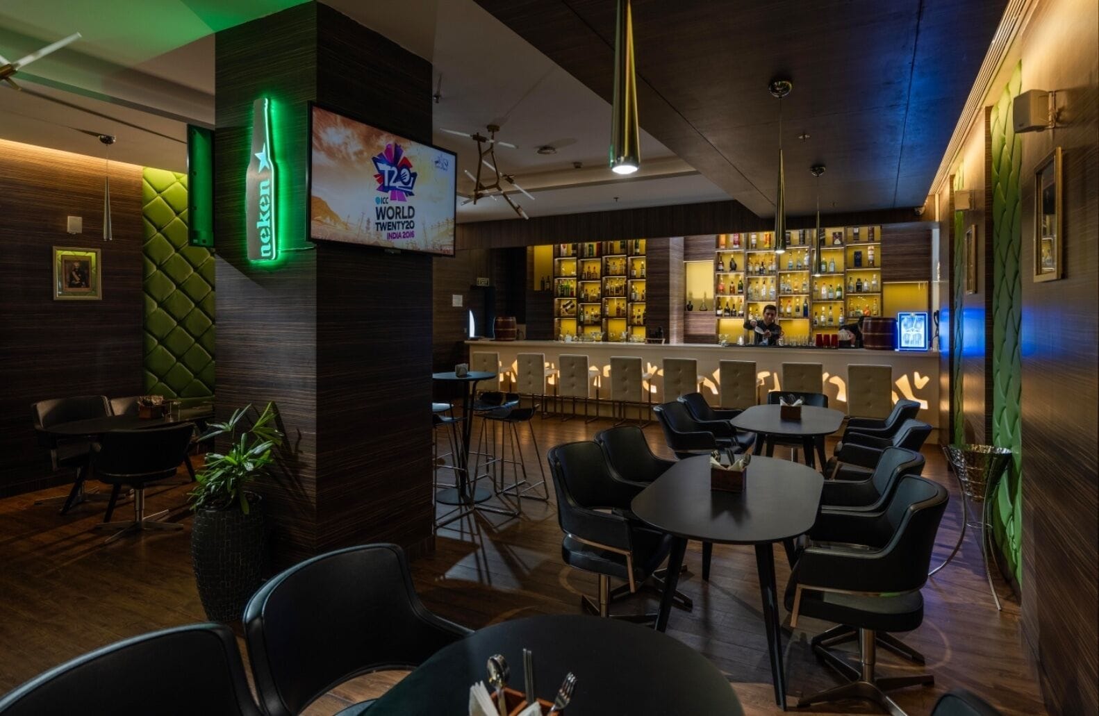 Una zona de bar con un letrero luminoso de T20 India 2016, un letrero de cerveza de neón y una barra provista de diversas botellas.