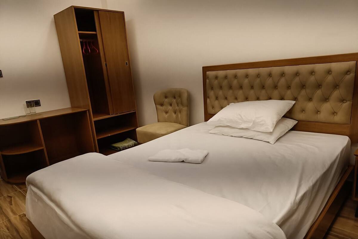 Ein ordentlich bezogenes Bett mit einer gepolsterten Kopfstütze, ein Holzschrank, ein kleiner Sessel und ein Nachttisch.