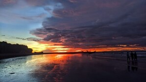 Ein Sonnenuntergang über einem Strand, mit Booten und silhouettierten Menschen vor dem Himmel.