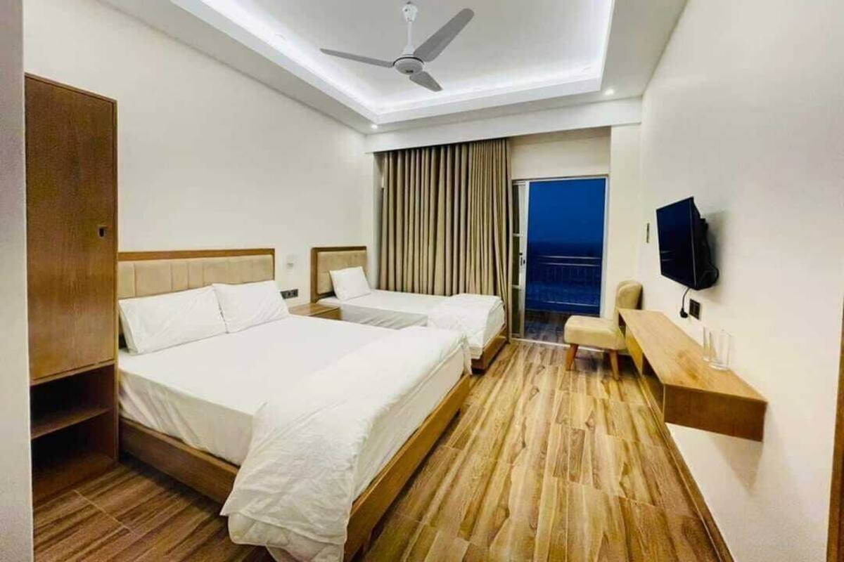 Ein Hotelzimmer mit zwei Betten, einem Deckenventilator, einem Fernseher, einer hölzernen Bank und einer Tür, die auf einen Balkon führt.