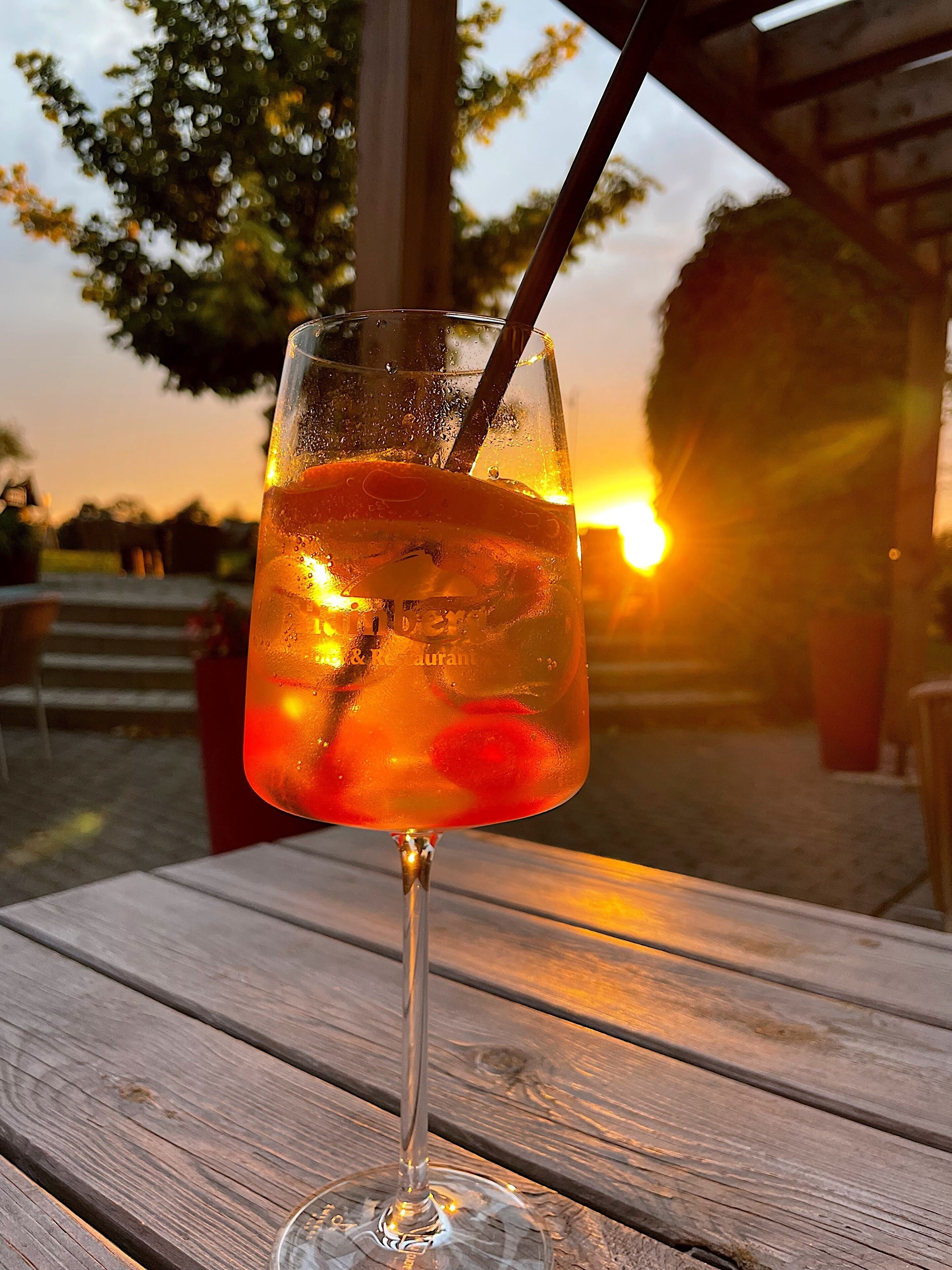 Ein Glas Eisgetränk mit Trinkhalm auf einem Holztisch, im Hintergrund ein Sonnenuntergang.
