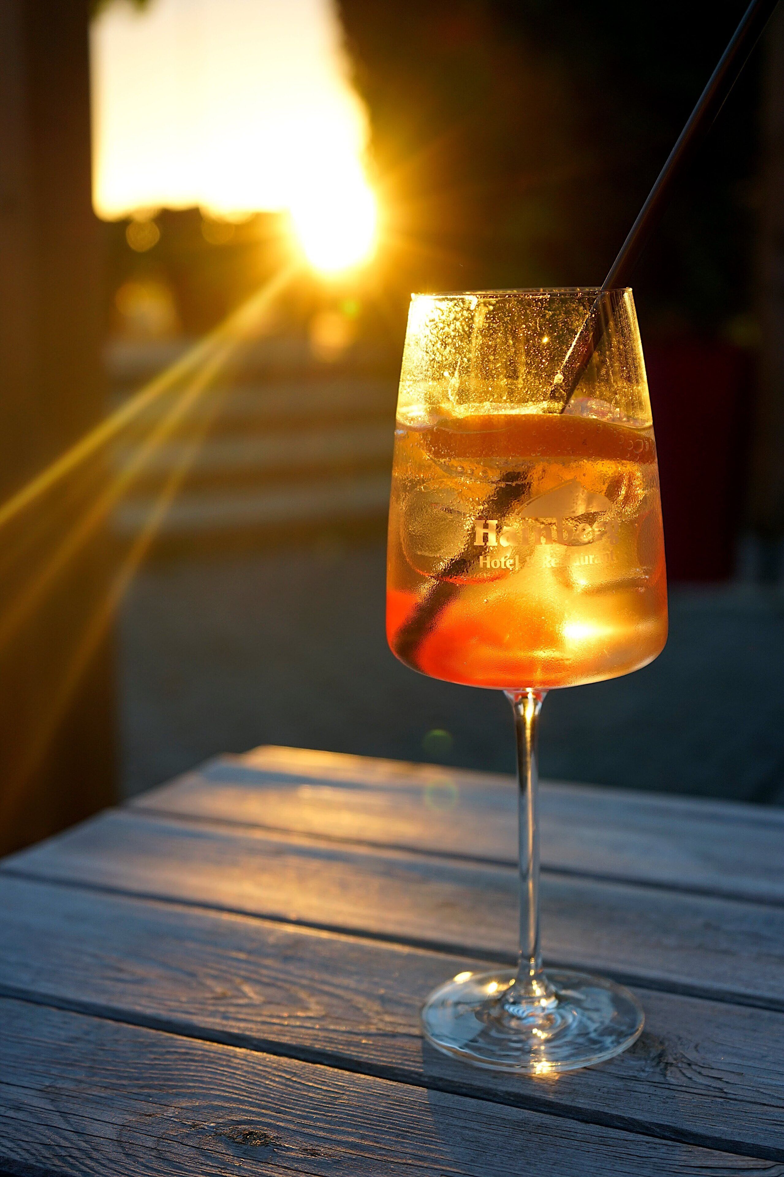 Ein Glas bernsteinfarbener Drink auf einem Holztisch, im Hintergrund unscharf ein helles Licht.