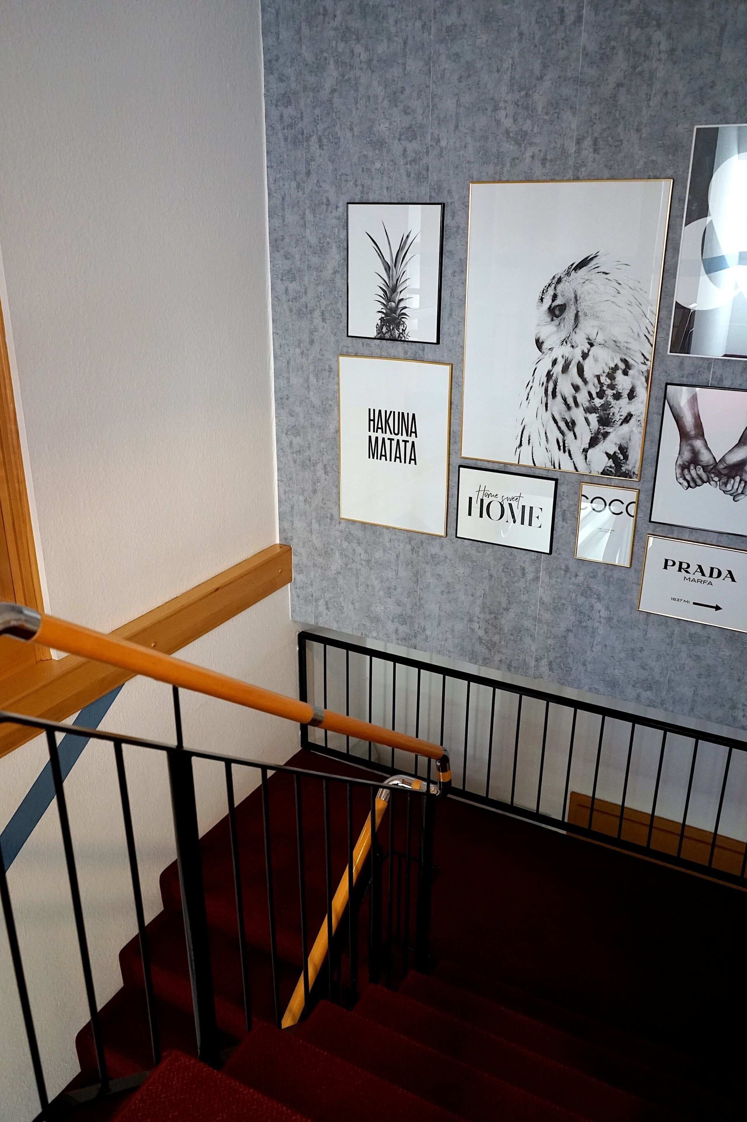 A staircase with a wooden handrail and black railings, leading to a landing with a grey textured wall adorned with framed artwork including a pineapple, an owl, and text phrases like "Hakuna Matata" and "Home".